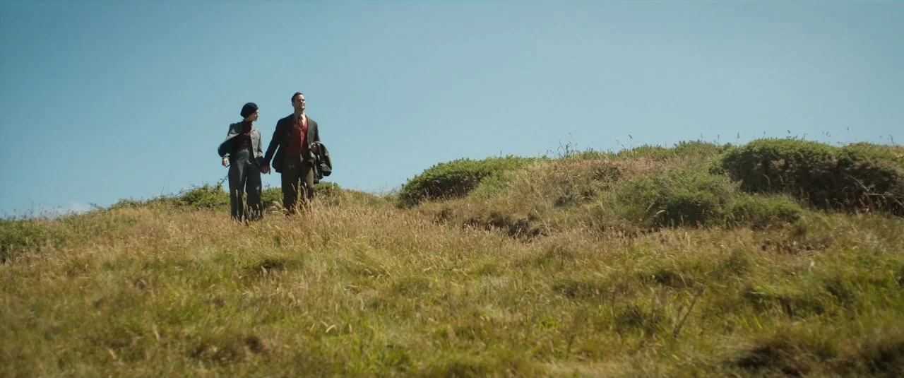 Rebecca movie still: wide — A man and woman walk together on a grassy, shrub-filled hillside under clear skies. They are holding hands while walking…