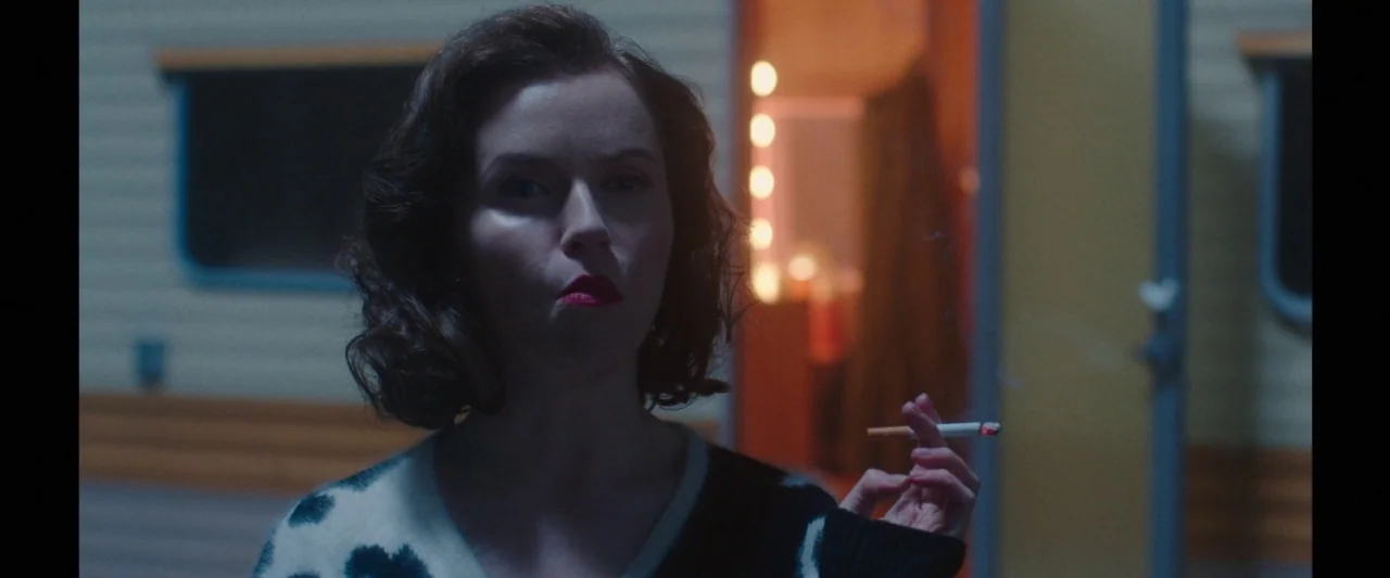 Censor (2021) movie still: medium — A woman stands in front of a trailer at night with dim lighting. She holds a lit cigarette while looking…