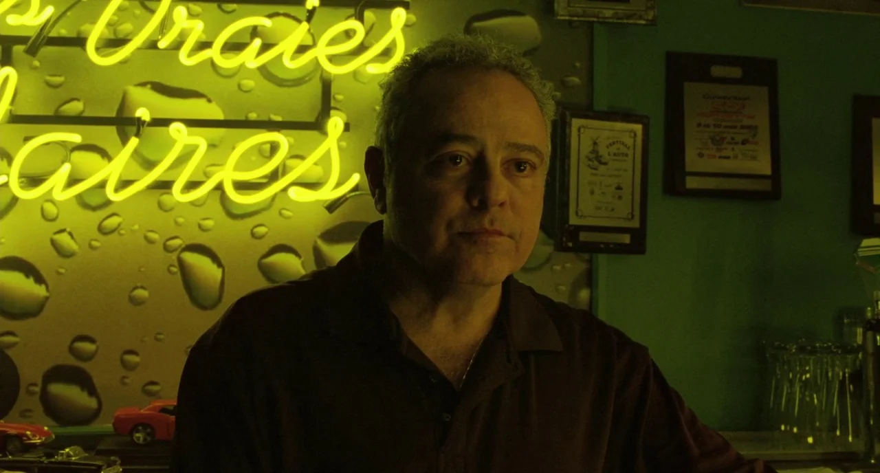 Tom at the Farm (2013) movie still: medium — A man stands before a wall featuring a bright yellow neon script sign. Framed certificates and small…