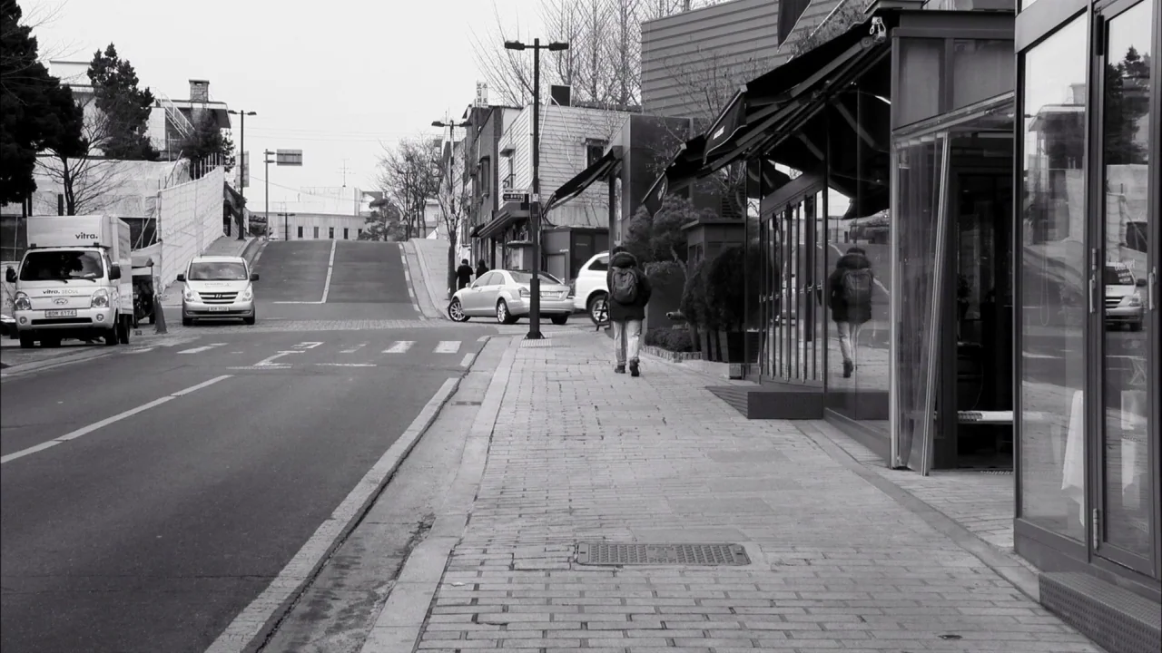 The Day He Arrives (2018) movie still: wide — An empty urban street scene with a pedestrian walking away on a brick sidewalk. Modern buildings with…
