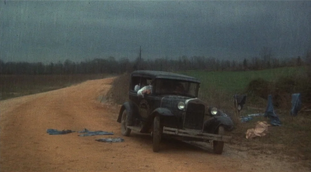 Thieves Like Us (1974) movie still: wide — A dark, vintage automobile is stationary on a rutted dirt road in a rural landscape. Pieces of blue…