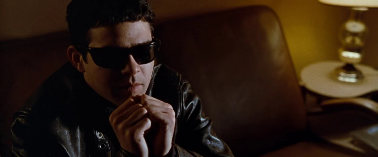 Empire Records (1995) movie still: medium — A man sits on a brown sofa inside a dimly lit room at night. He is clasping his hands together beneath…