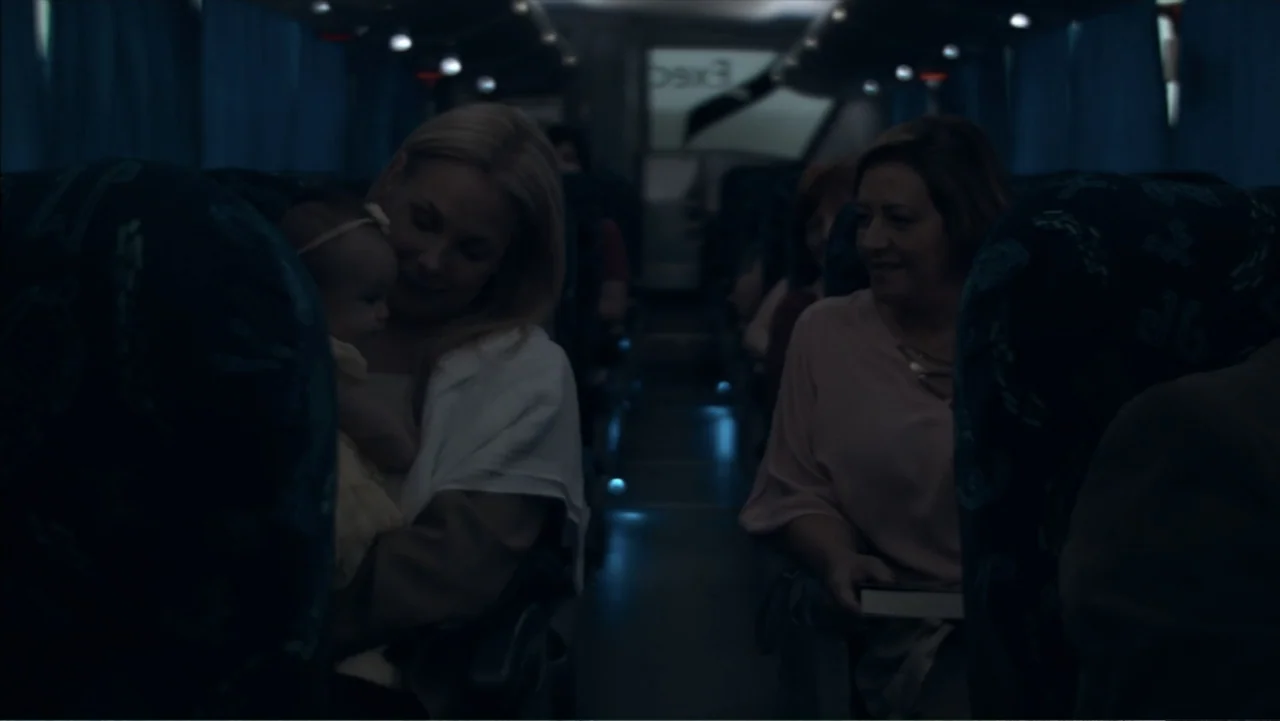 The Grotto (2022) movie still: medium — The interior cabin of a bus at night features rows of patterned seats. One woman holds a small baby while…