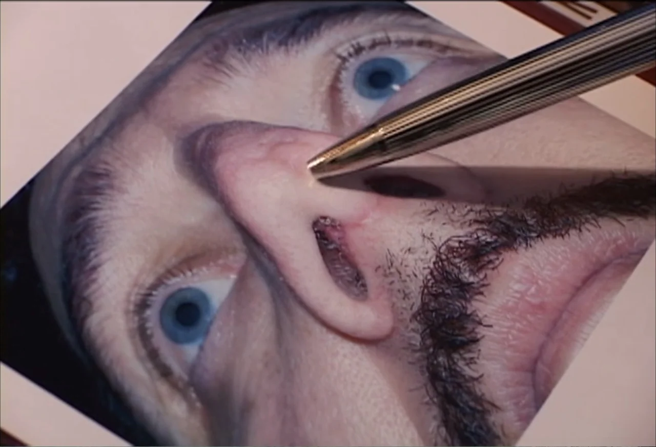 Plastic Disasters (2006) movie still: closeup — A close-up view of a printed photograph held on a surface. A metallic pen rests against the image,…
