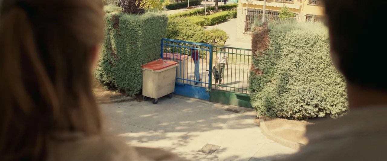 Voy a pasármelo bien (2022) movie still: wide — A sunny outdoor courtyard features a blue metal gate flanked by tall pruned hedges. Two youths walk…