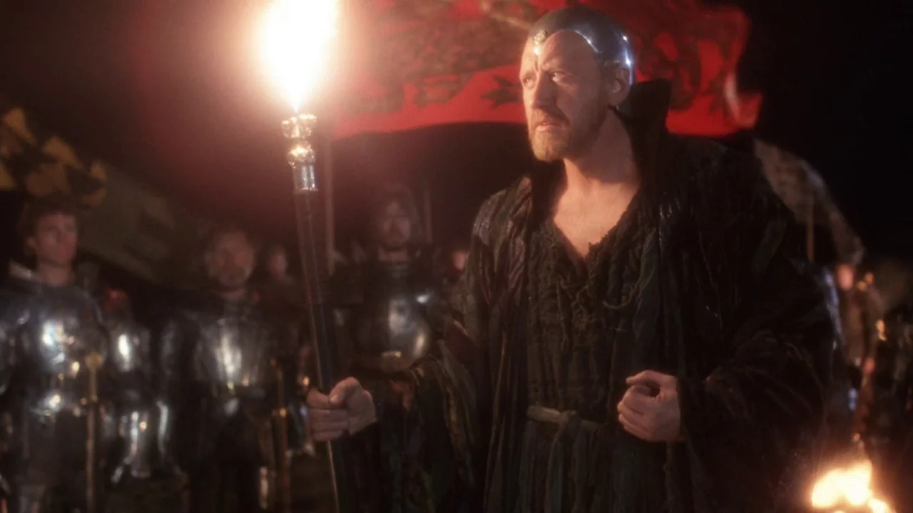 Excalibur (1981) movie still: medium — A nighttime military camp at night with soldiers in metal armor visible in the back. A bearded man in a…