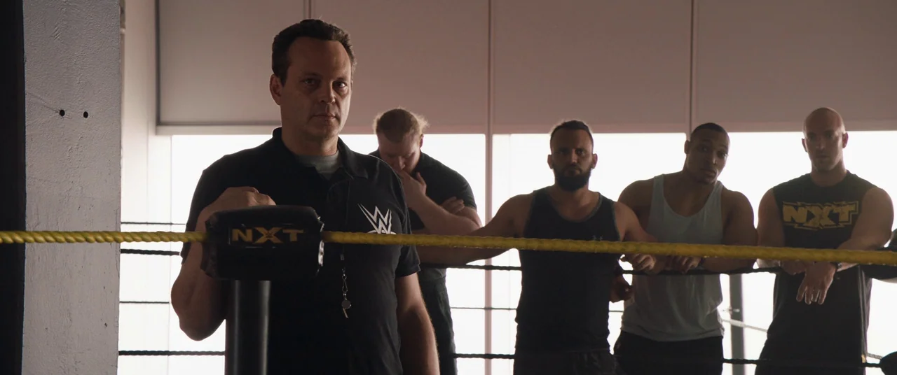 Fighting With My Family (2019) movie still: medium — A brightly lit training space features a professional wrestling ring with yellow ropes. Five…