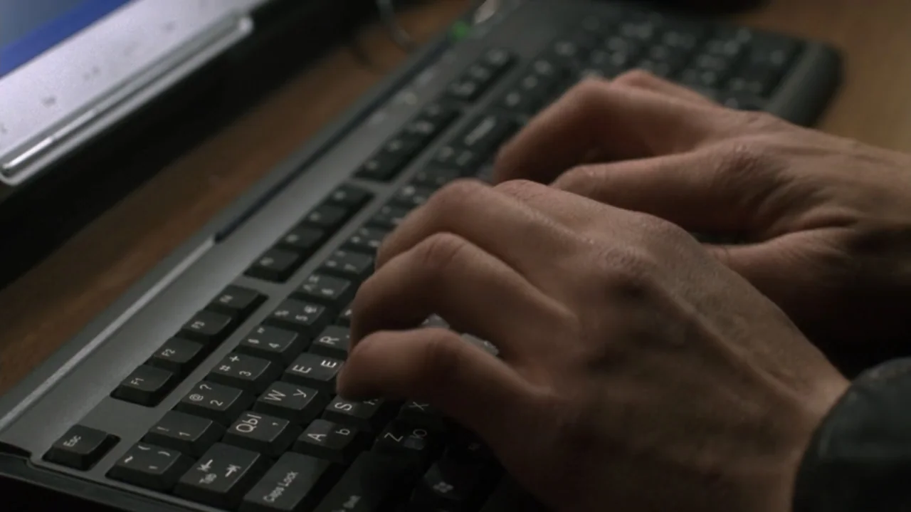 The Shepherd (2023) movie still: closeup — A close view of human hands resting on a black computer keyboard surface. The hands are engaged in typing…