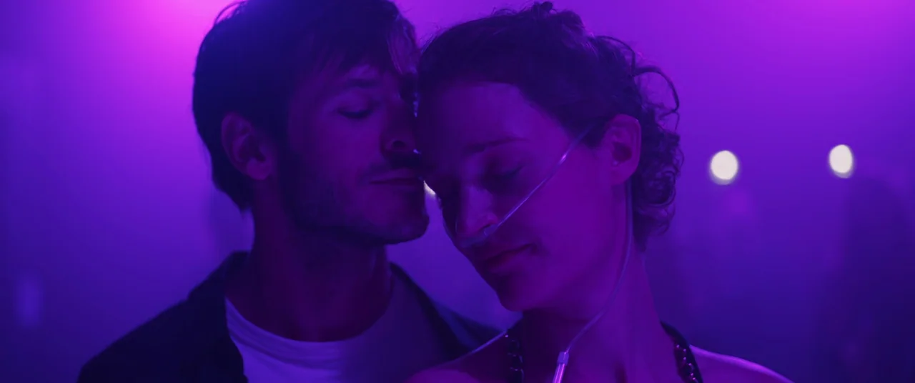 More Than Ever (2022) movie still: closeup — A dark, purple-lit venue filled with people moving in the blurry background. A man and a woman stand…