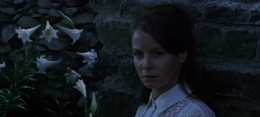 Ryan’s Daughter (1970) movie still: closeup — A woman stands against a dark, rough-hewn stone wall next to white lily flowers. She is positioned in…