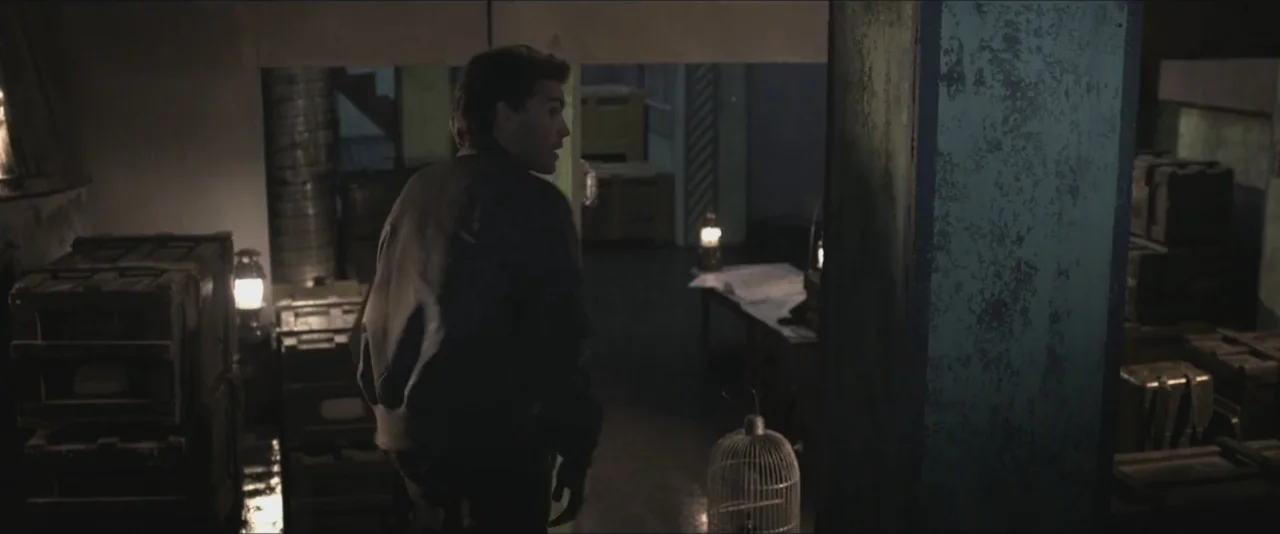 Darkest Hour (2017) movie still: medium — A dark, cluttered storage room filled with wooden crates is lit by small oil lanterns. A young man wearing…