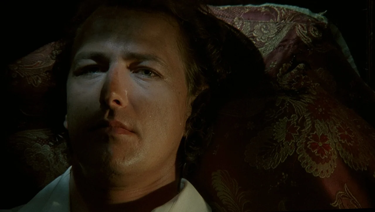 Necronomicon: Book of Dead (1993) movie still: closeup — A man lies with his head resting on a dark red ornamental pillow. He looks forward while…