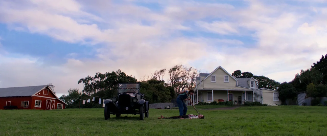 Pearl (2022) movie still: wide — A rural farmstead features a yellow house, a red barn, and a grassy lawn. One person stands over a prone individual…