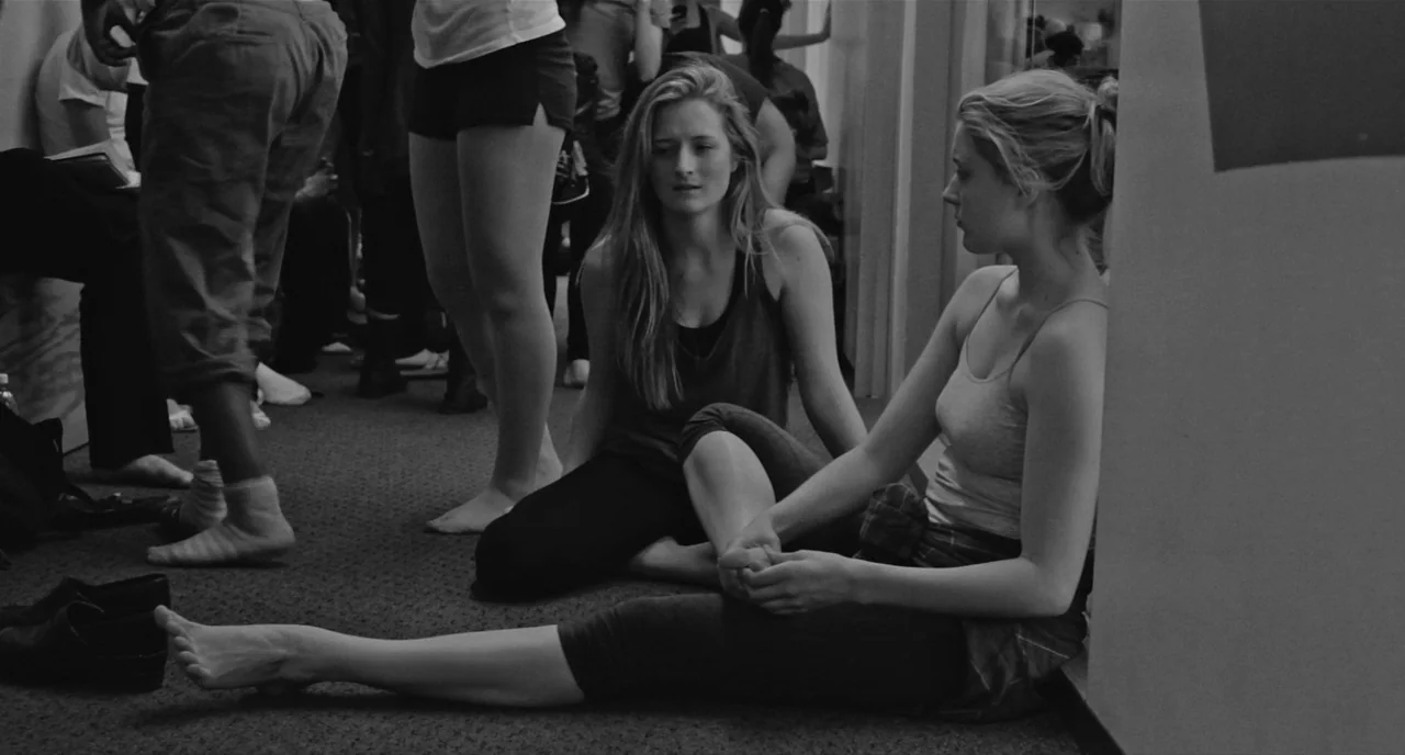 Frances Ha (2012) movie still: medium — Two young women sit on the floor of a crowded, carpeted practice space. One woman holds her foot while…