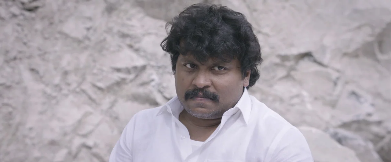 Peranbu (2019) movie still: medium — An adult male stands in front of a textured, light-colored rocky outcropping outdoors. He faces the camera…