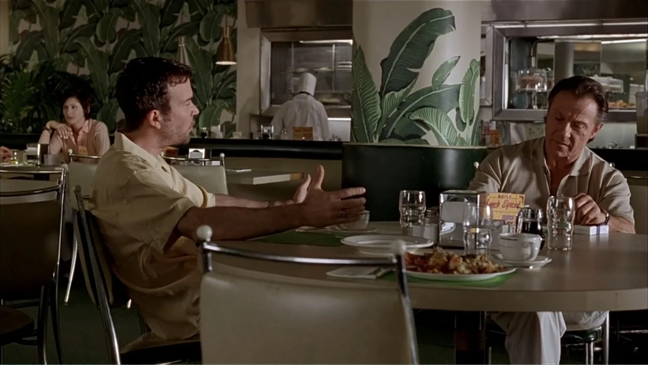 Ajuste de Cuentas (2016) movie still: medium — A 1990s-style diner interior features banana leaf wallpaper and white laminate furniture. Two men sit…