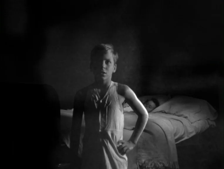 The Night Of The Hunter (1955) movie still: medium — A dark, sparsely furnished room contains a bed where another person lies sleeping. A young boy…