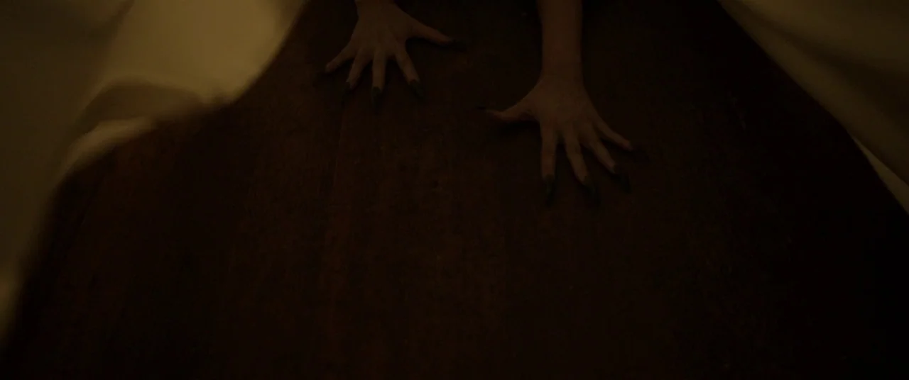 Satan’s Slaves (2017) movie still: closeup — A dark wooden floor serves as the surface for two hands with pointed nails. The hands rest on the wood…