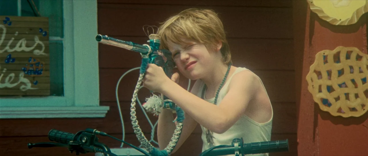 Riddle of Fire (2023) movie still: medium — A child sits on a bicycle in front of a red wooden wall. The boy aims a modified toy gun while squinting…