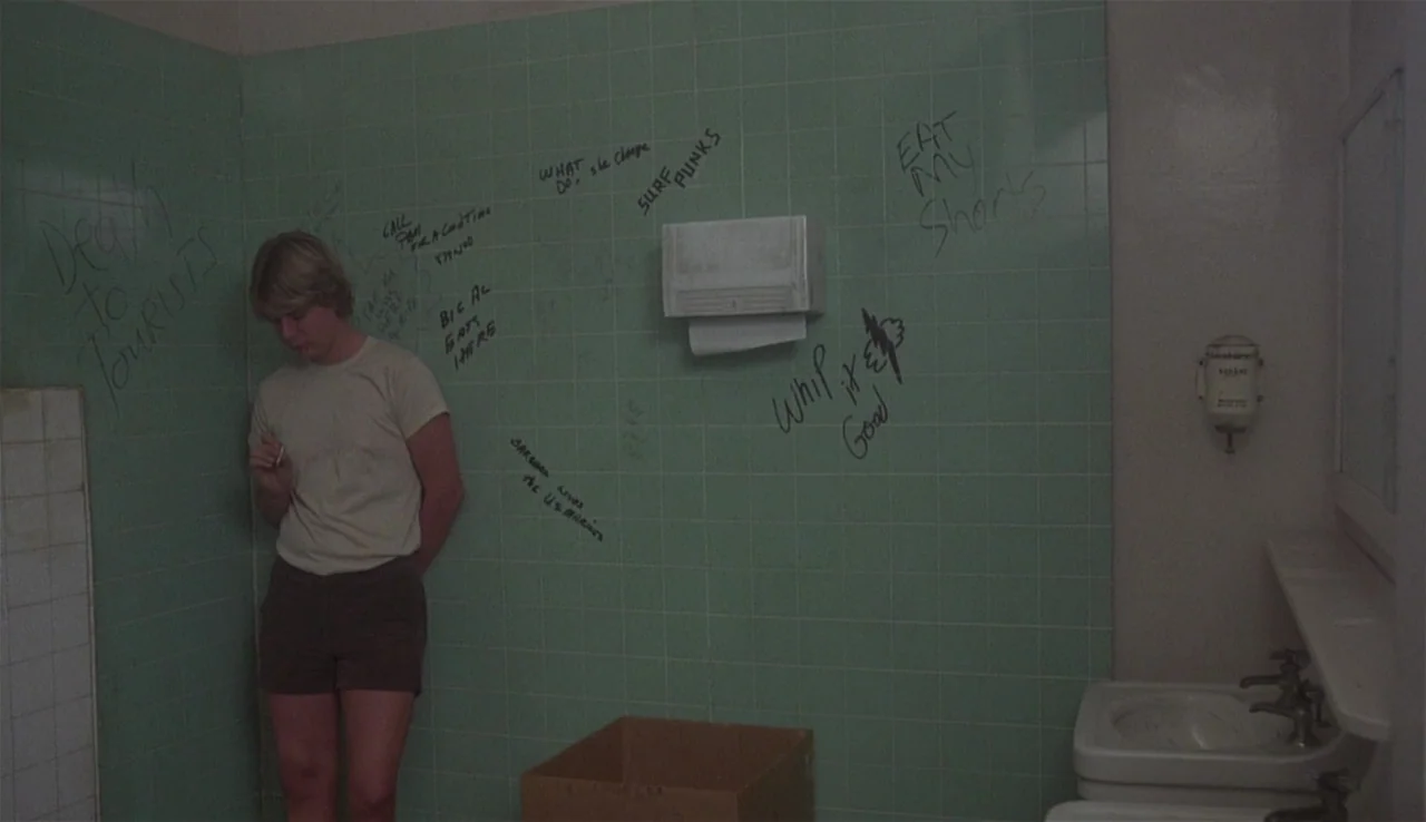 Body Heat (1981) movie still: medium — A utilitarian public restroom features mint green wall tiles covered in black marker graffiti. A teenage boy…