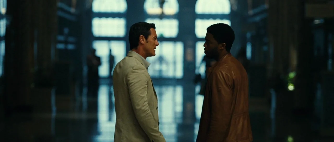 Message From The King (2016) movie still: medium — A spacious, dimly lit building interior with a row of arched windows in background. Two men in…