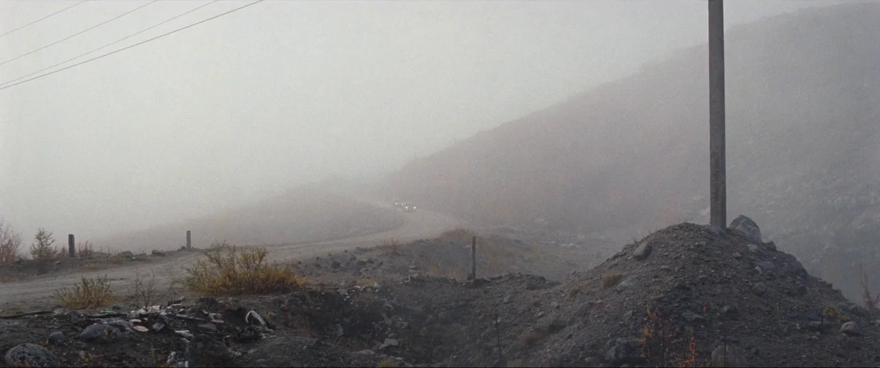 Leviathan (2014) movie still: wide — A desolate, foggy landscape features a winding gravel road amidst rocky, barren terrain. A lone vehicle travels…