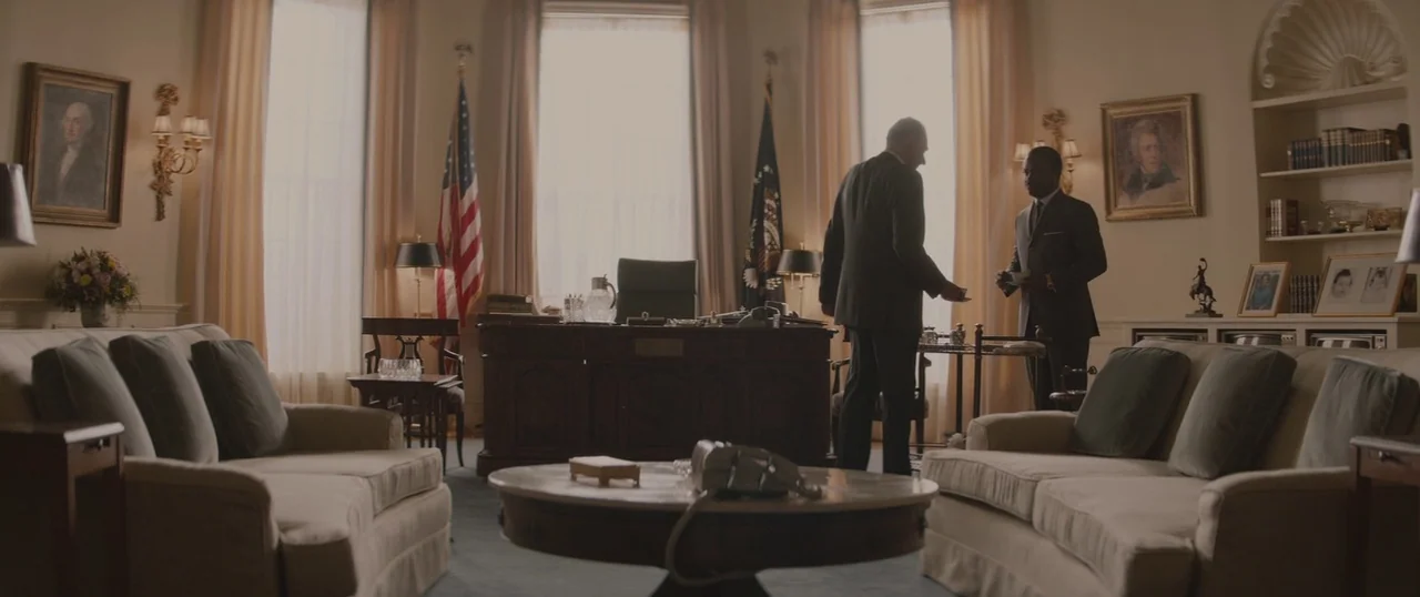 Selma (2014) movie still: wide — An oval-shaped administrative room features two light-colored sofas, a large wooden desk, and flags. Two men in…