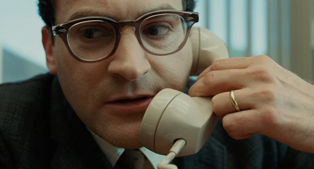 A Serious Man (2009) movie still: closeup — An office environment with vertical blinds visible in the out-of-focus background. A man holds a rotary…
