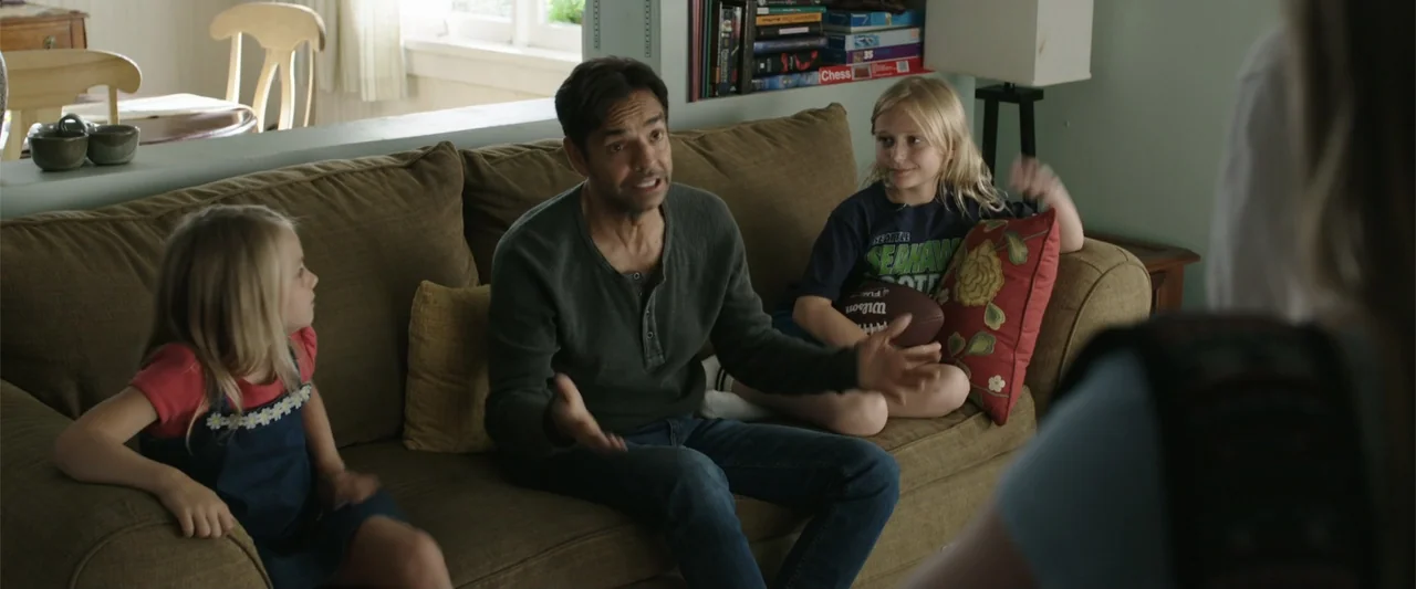 Overboard (2018) movie still: medium — A living room interior with an olive couch and shelves filled with books. A man gestures while talking to two…