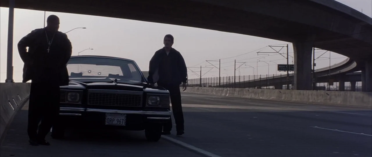 Training Day (2001) movie still: wide — A wide paved highway stretches beneath a massive concrete overpass in an urban setting. Two men stand beside…