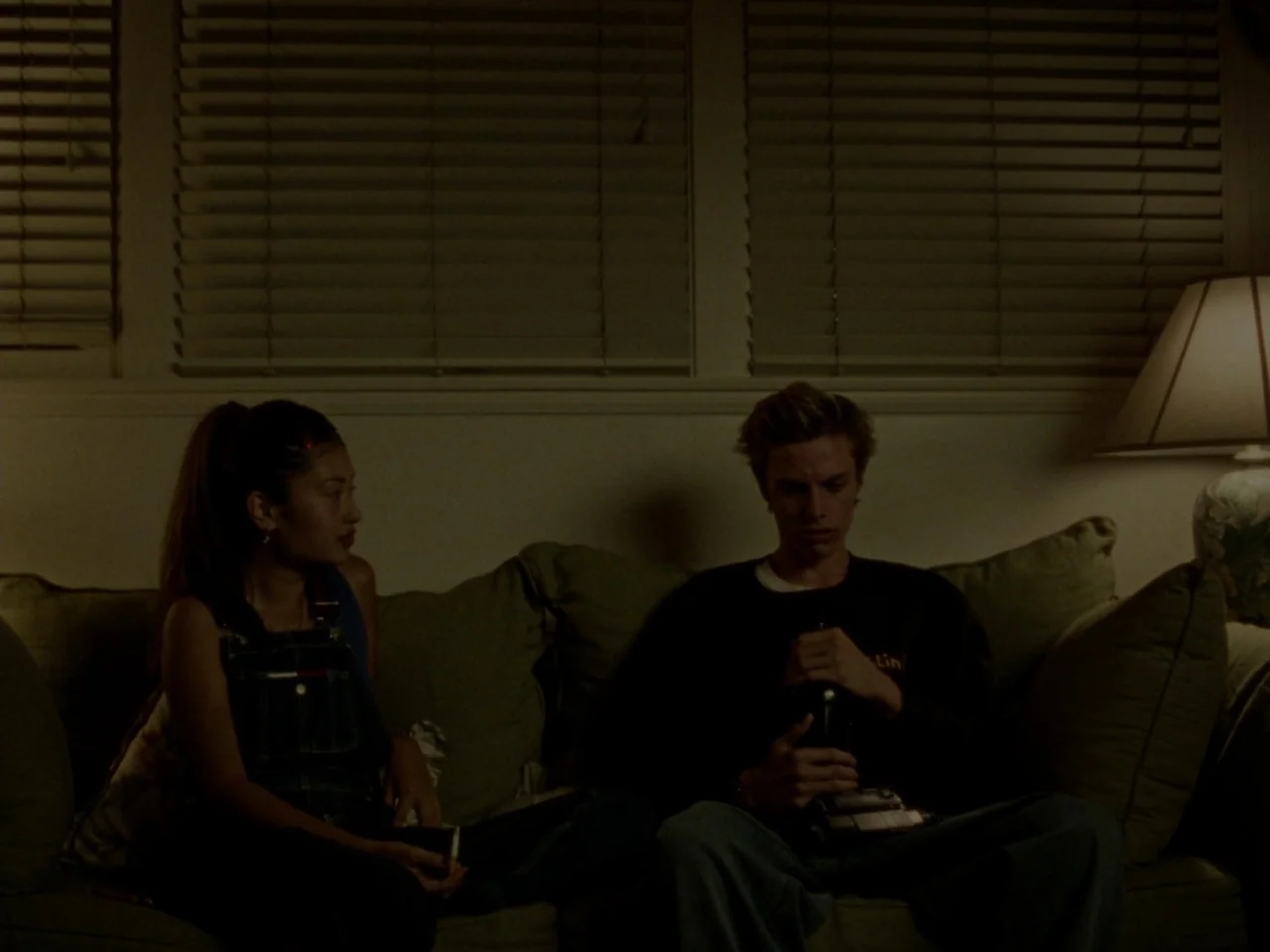 Mid90s (2018) movie still: medium — A dimly lit room features a pair of individuals seated on a green couch. The man holds a camcorder in his lap…