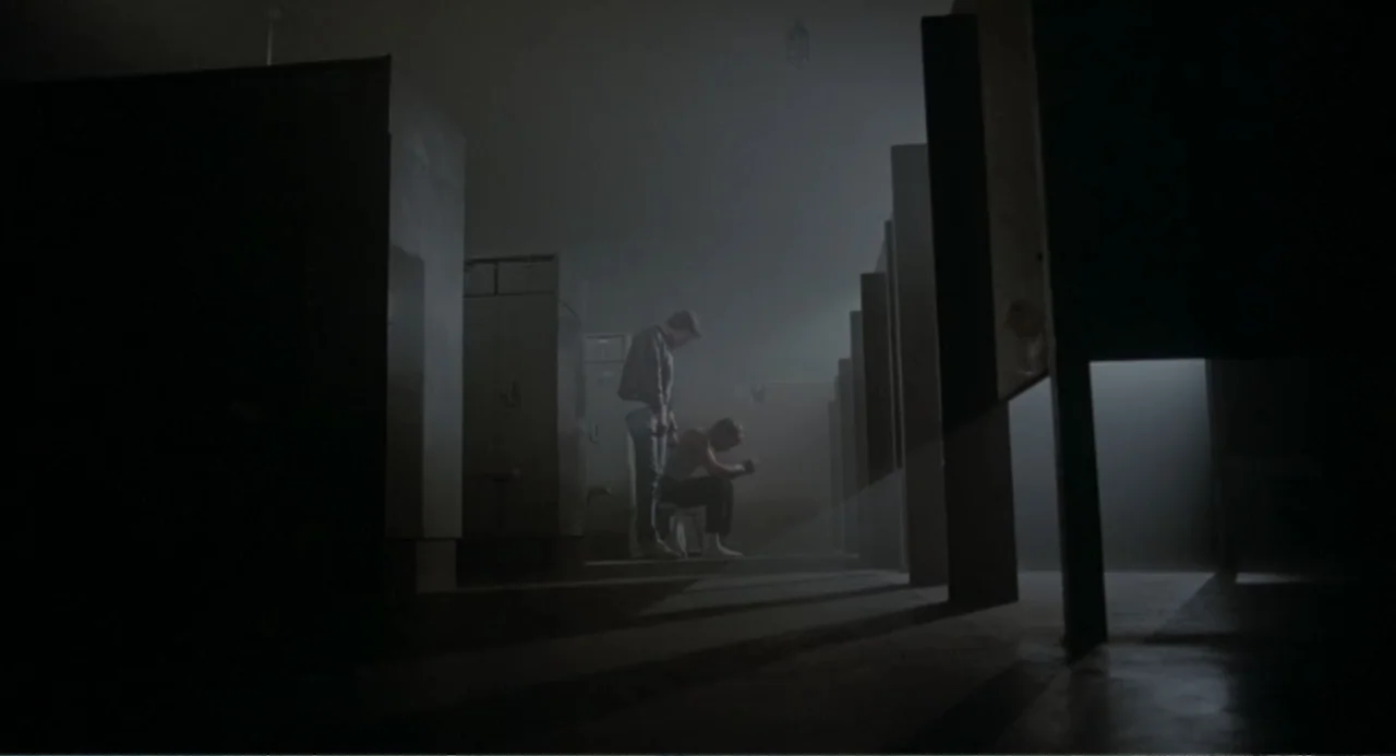Dangerously Close (1986) movie still: wide — A dark locker room contains rows of metallic cabinets and benches under harsh light. One man stands…