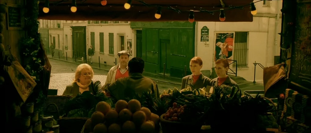 Amelie (2001) movie still: wide — An outdoor market stall filled with produce overlooks a quiet cobblestone street in Paris. A small group of people…