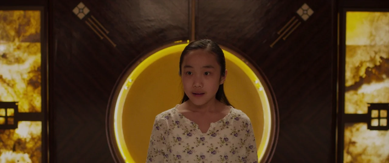 The Perfection (2018) movie still: medium — An interior space features a dark wood wall with a central circular glowing light. A young person stands…