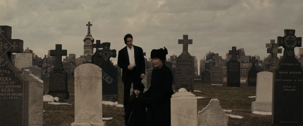 Winter's Tale (2014) movie still: fullbody — A historic cemetery with rows of weathered stone markers under a cloudy sky. A man in formal attire…