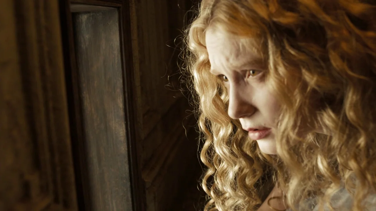 Alice in Wonderland (2010) movie still: closeup — A dimly lit room with dark wood wall paneling. A young woman with curly blonde hair gazes toward…