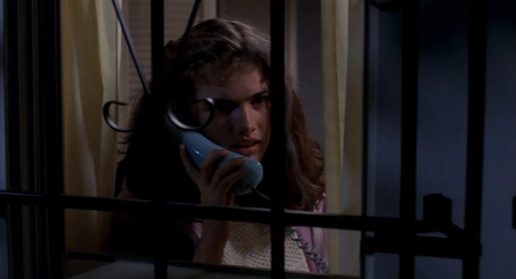A Nightmare On Elm Street (1984) movie still: closeup — A view through a black metal security gate into a dimly lit room. A young woman holds a…