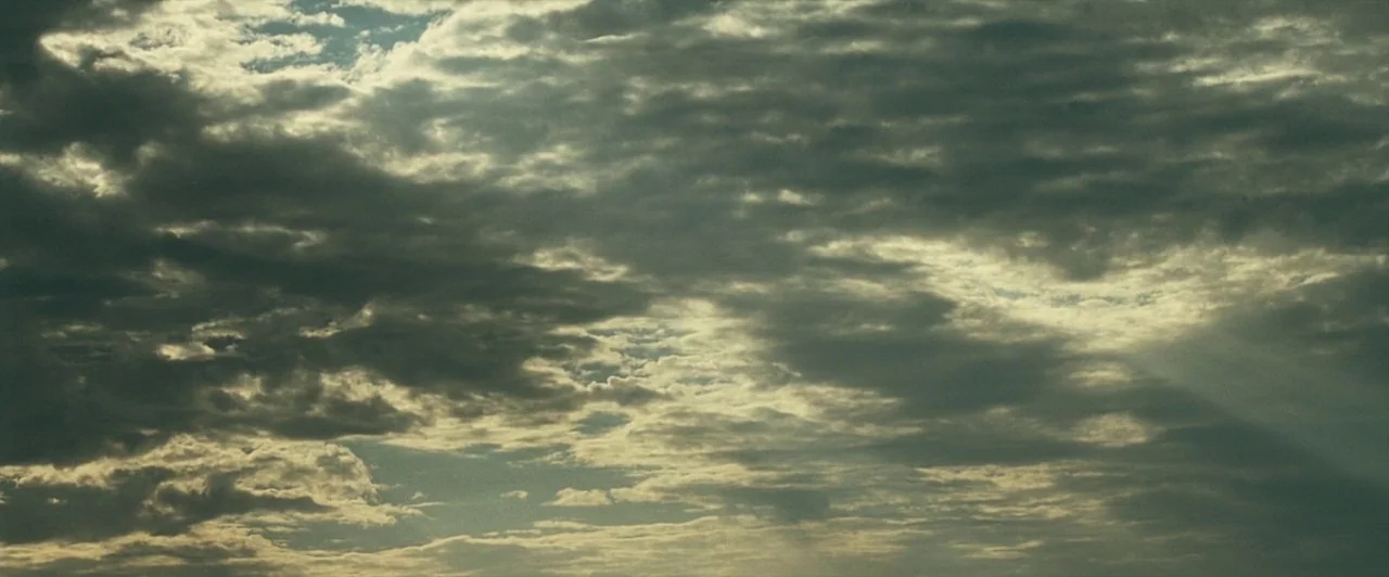 The Proposition (2005) movie still: establishing — A wide view of the sky filled with dense, layered cloud formations. Patches of light break…