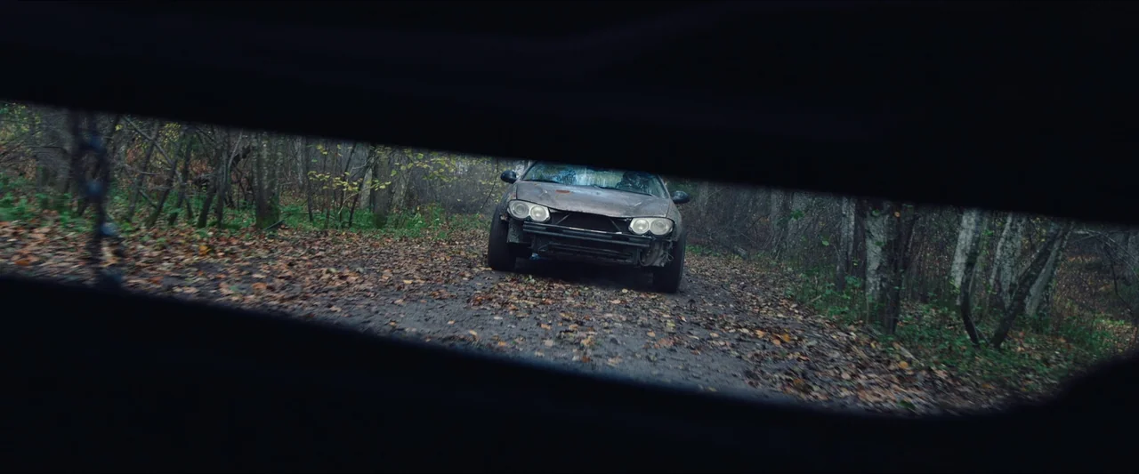 Azrael (2024) movie still: wide — A damaged sedan is parked on a leaf-strewn dirt road within a dense forest. The car lacks a front bumper and…