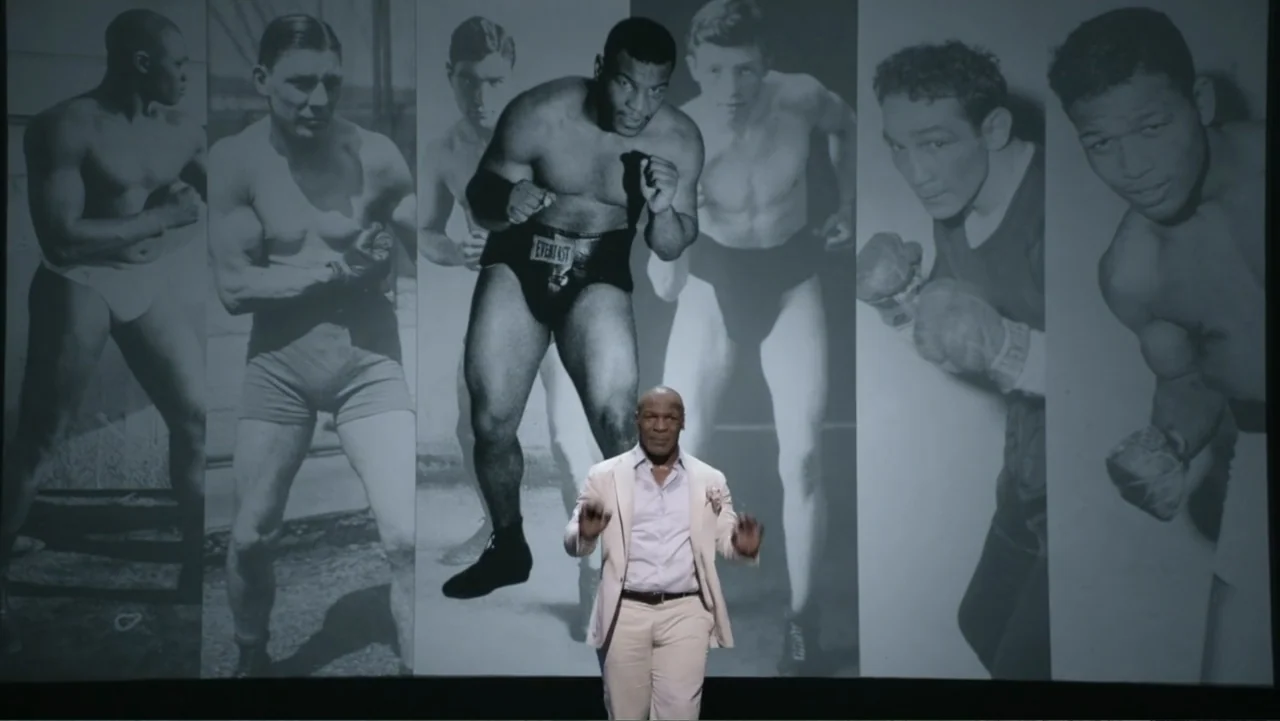 Mike Tyson: Undisputed Truth (2013) movie still: medium — A man stands on a stage against a wall covered in large vintage boxing photographs. The…