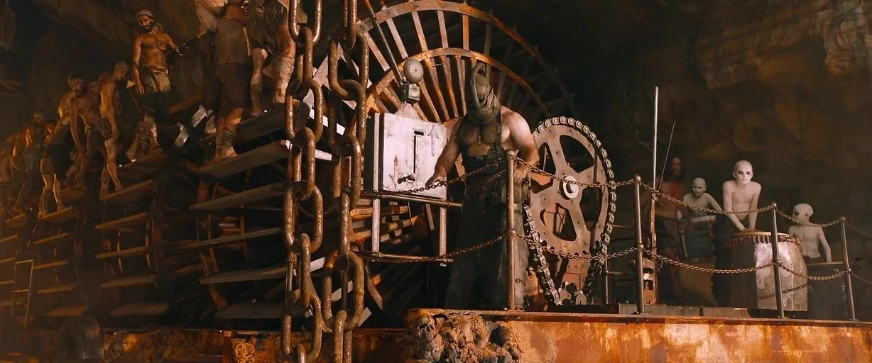 Mad Max: Fury Road (2015) movie still: wide — A dark, cavernous space is filled with heavy, rusted, complex iron machinery and chain systems. A…