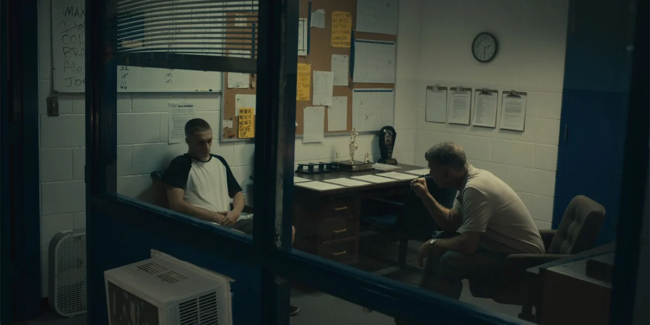 All Roads to Pearla (2019) movie still: medium — A small office with white cinderblock walls features a desk, corkboard, and clipboards. An older…