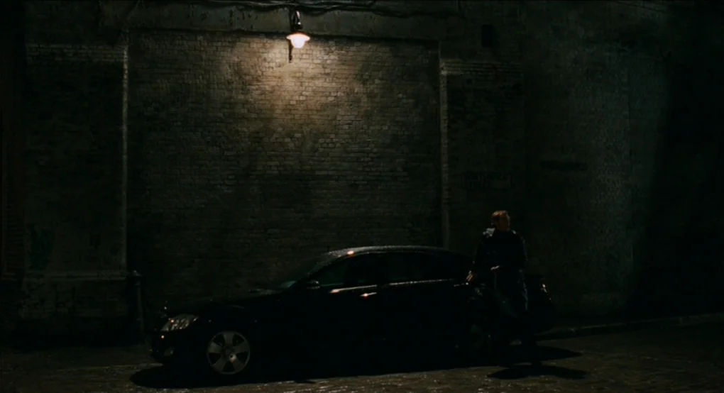 Eastern Promises (2007) movie still: wide — A dark brick wall lines a cobblestone street under the light of a streetlamp. A man stands next to a…
