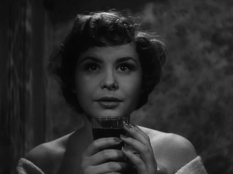 Waiting Women (1952) movie still: closeup — A woman stands in a dimly lit room with textured wallpaper behind her. She holds a small glass in both…