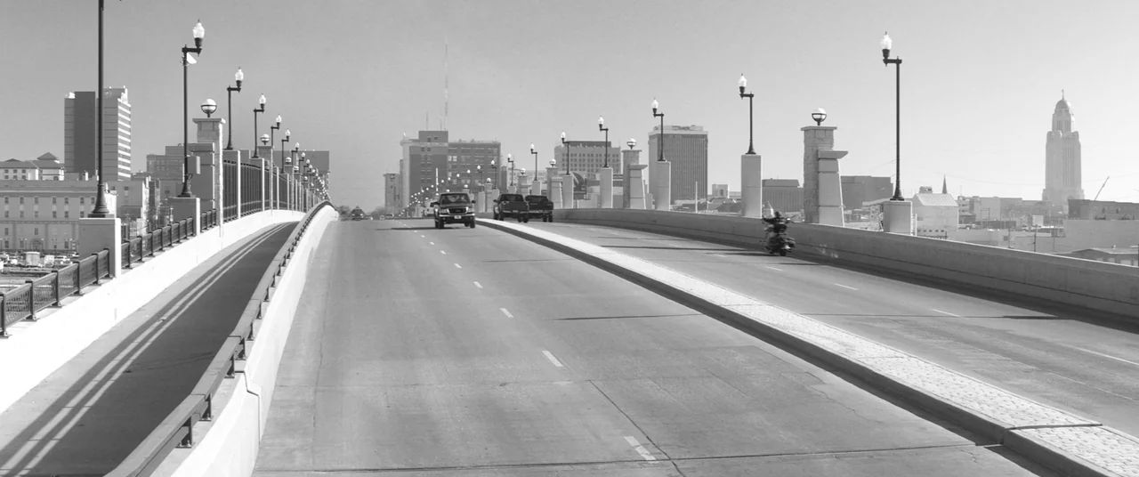 Nebraska (2013) movie still: establishing — A wide concrete bridge spans over a city, lined with decorative period-style lamp posts. Several cars…
