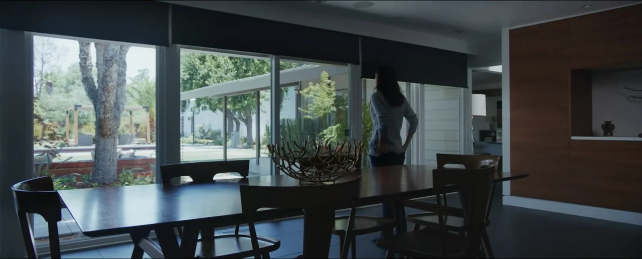 Hostages (2017) movie still: medium — A modern dining room features a dark wood table and chairs near large glass doors. A woman stands by the…