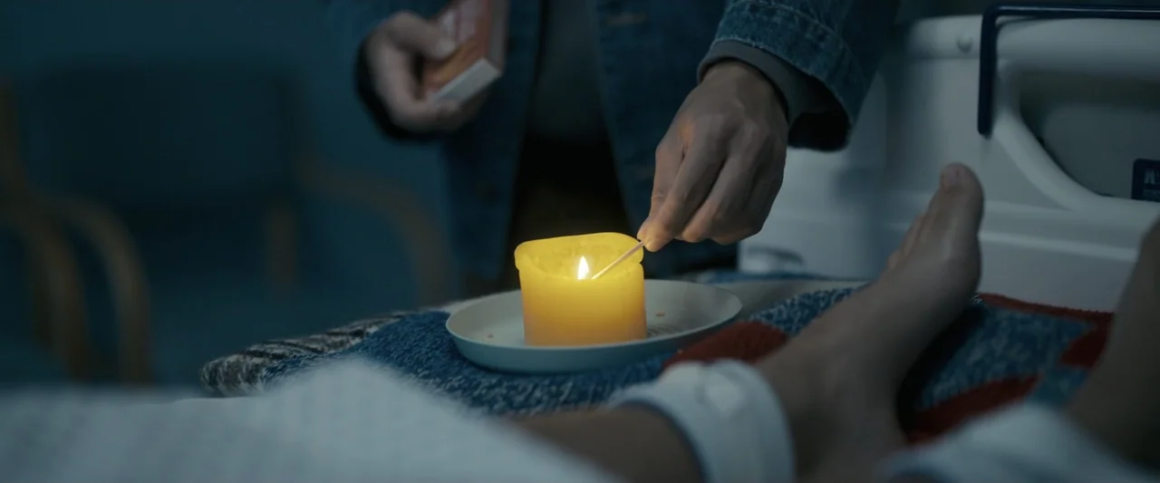 Talk to Me (2023) movie still: medium — A dimly lit hospital room features a medical bed with a patient's feet visible. A person in a denim jacket…