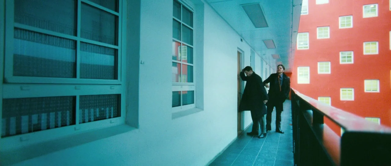 Filth (2013) movie still: wide — A modern outdoor residential walkway features a stark white wall and a bright red building. Two men stand together…