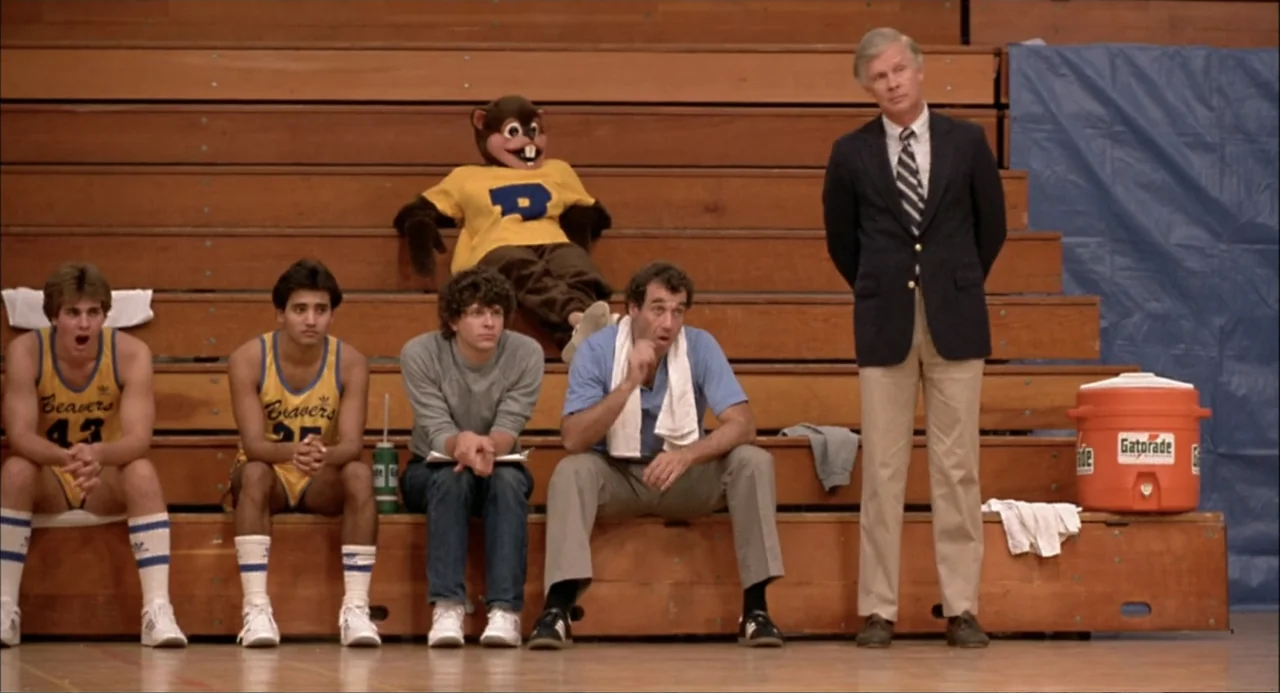 Teen Wolf (1985) movie still: fullbody — A group of five men sits or stands on wooden bleachers inside a gym. A person wearing a beaver mascot…