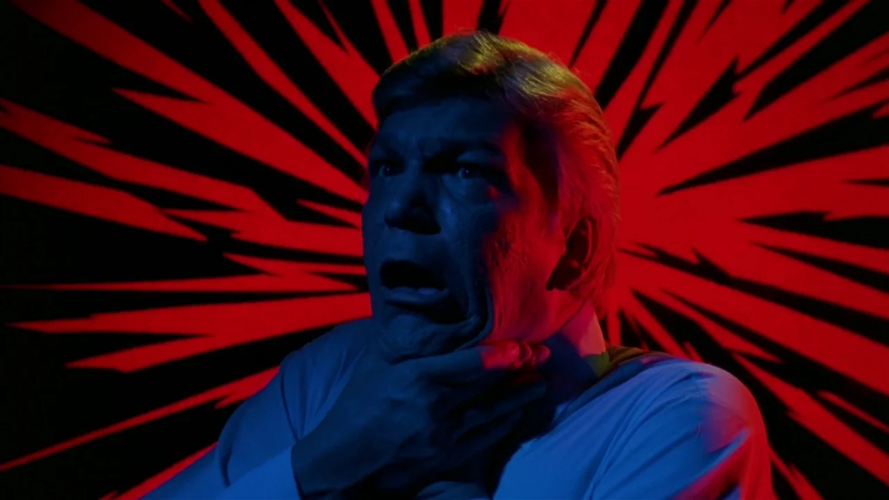 Creepshow (1982) movie still: closeup — A man stands against a graphic backdrop of radiating red and black lines. He grips his own throat with both…